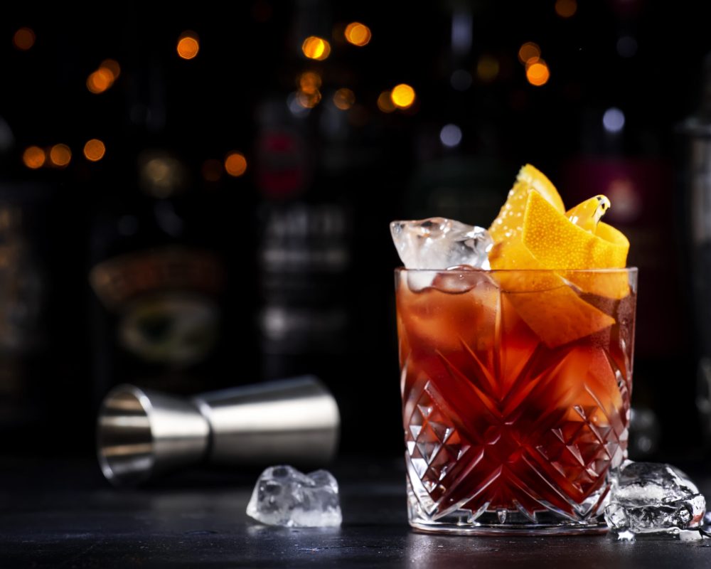 Espresso Negroni alcoholic cocktail drink with dry gin, red vermouth and red bitter, espresso and coffee liqueur, orange slice and ice cubes. Black bar counter background, bar tools, bottles